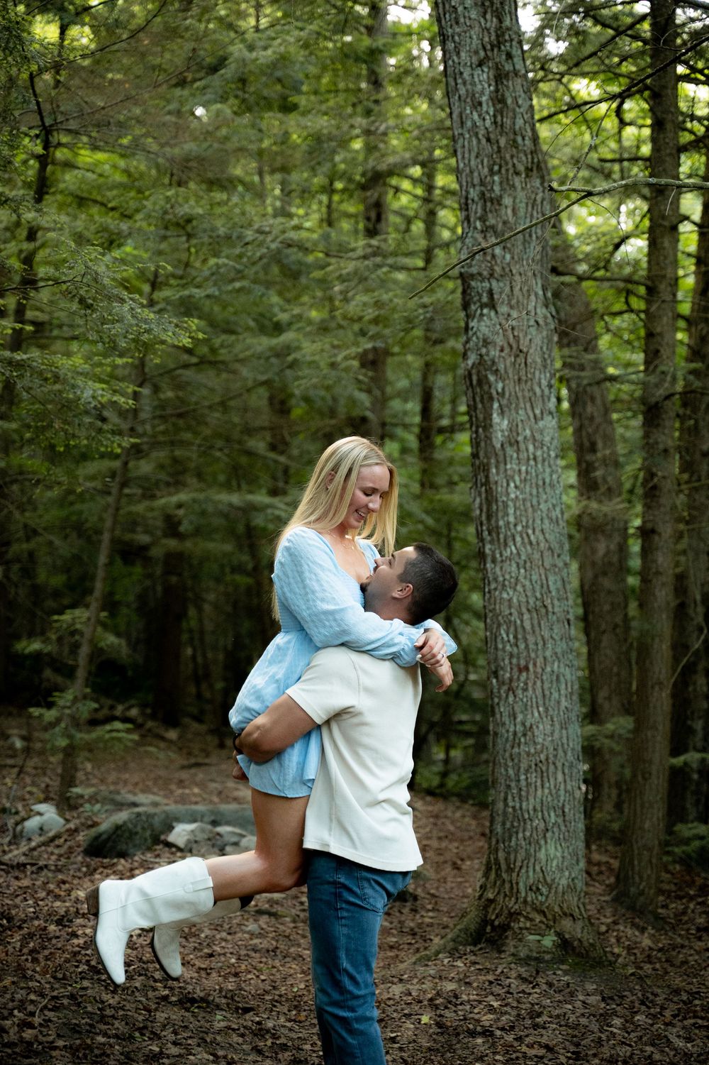 A man lifts a smiling blonde woman in a forest, both gazing at each other lovingly among tall green trees.
