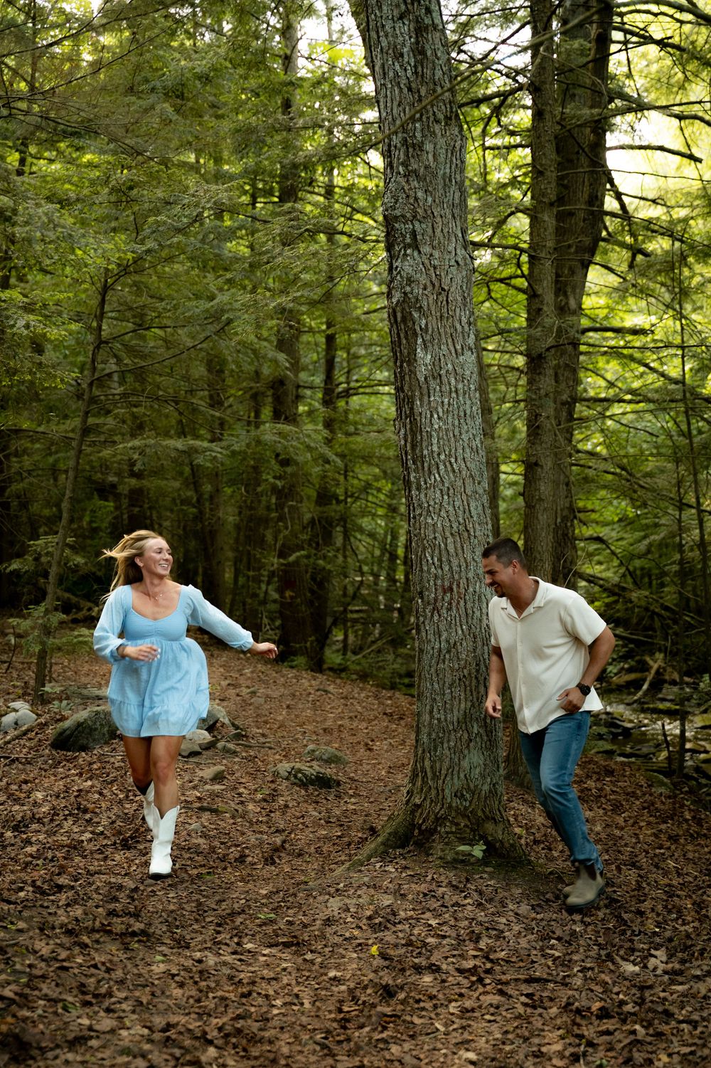 Couple playfully running through a lush green forest, woman in blue dress and white boots, man in jeans and cream shirt.