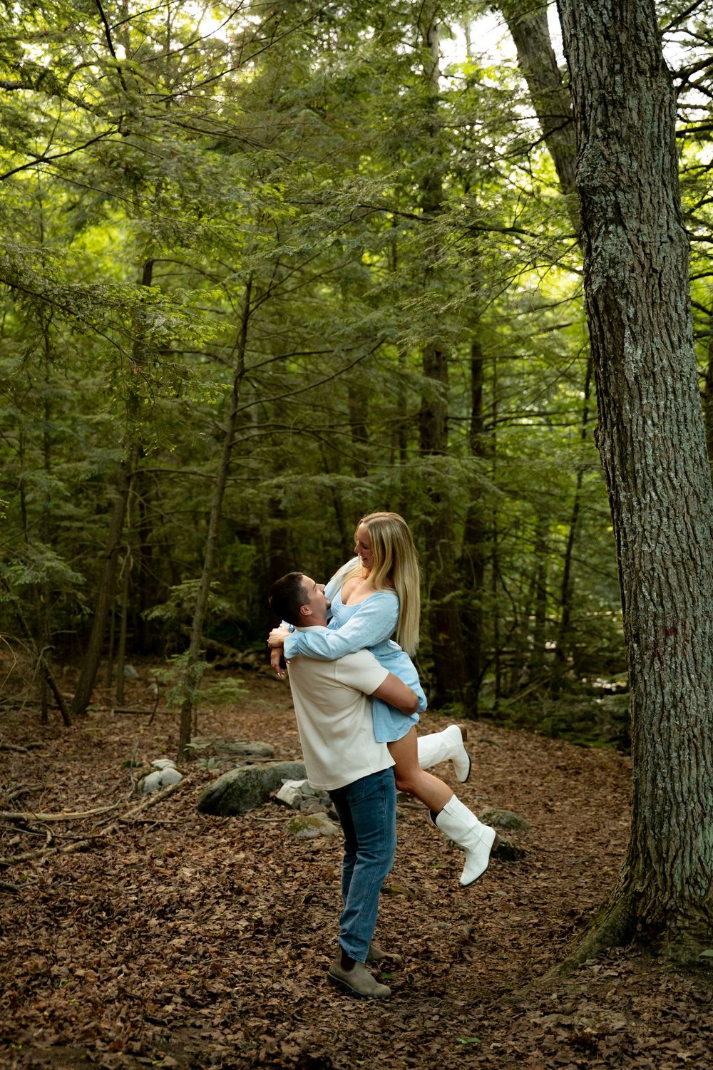 A man lifts a woman in a romantic embrace among lush green trees on a forest trail covered in fallen leaves.