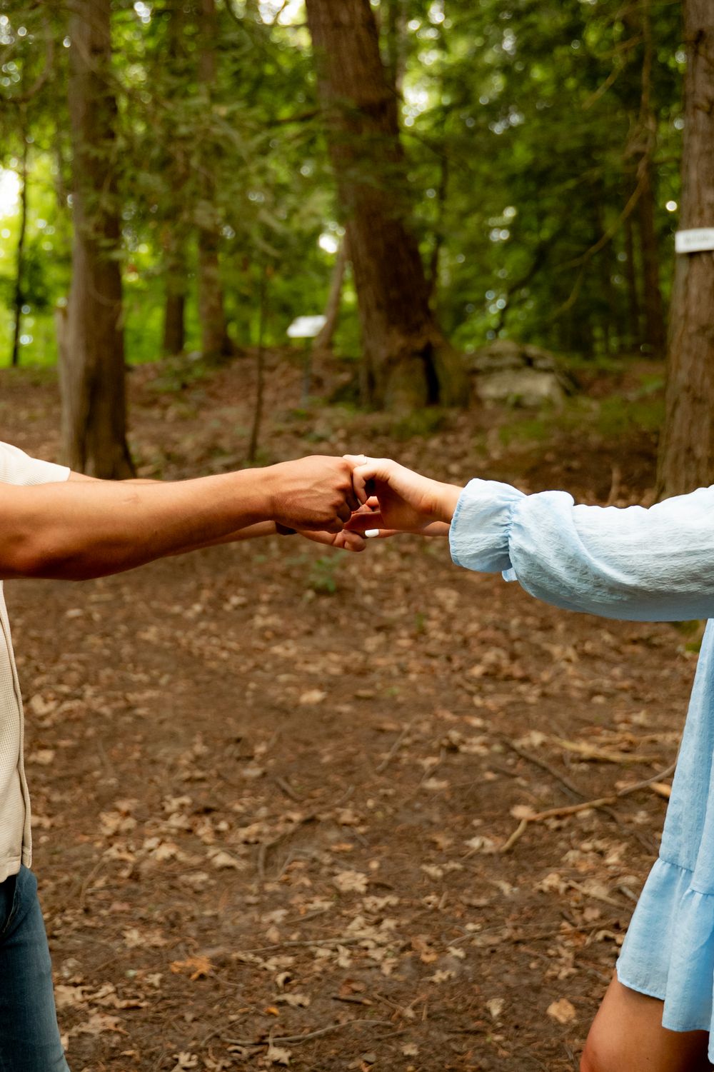 Two people fist bumping in a lush green forest setting, one wearing a light blue dress.