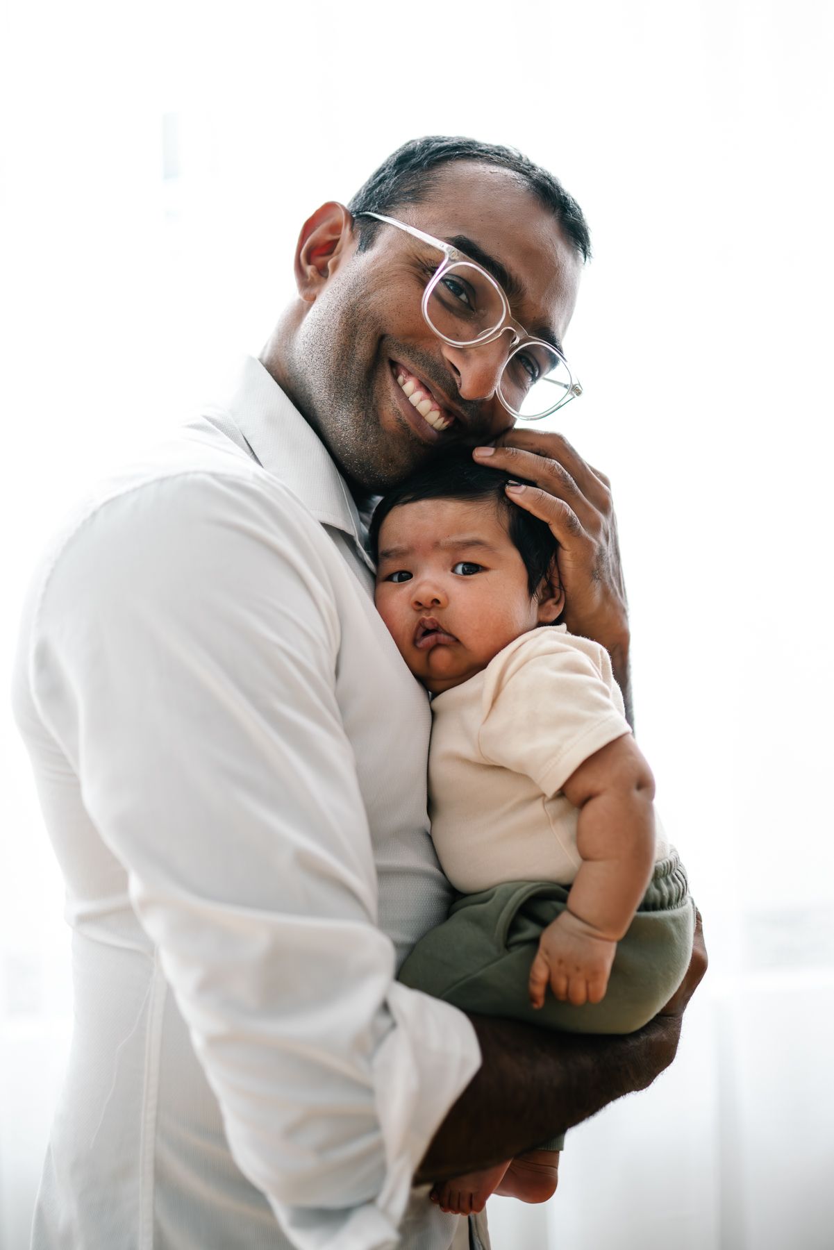Smiling man with glasses holding a cute baby in a white onesie, sharing a tender moment together during a Melbourne in-home Newborn Session