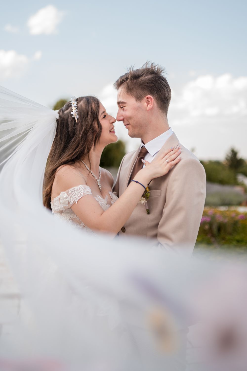Bride and groom share a romantic moment, nearly kissing outdoors, with veil flowing in the breeze on their wedding day.