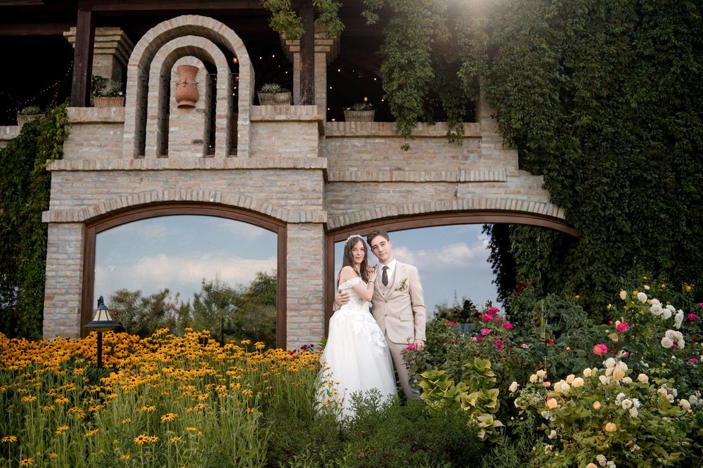 Bride and groom pose in lush garden with colorful flowers beside elegant stone building on sunny wedding day.
