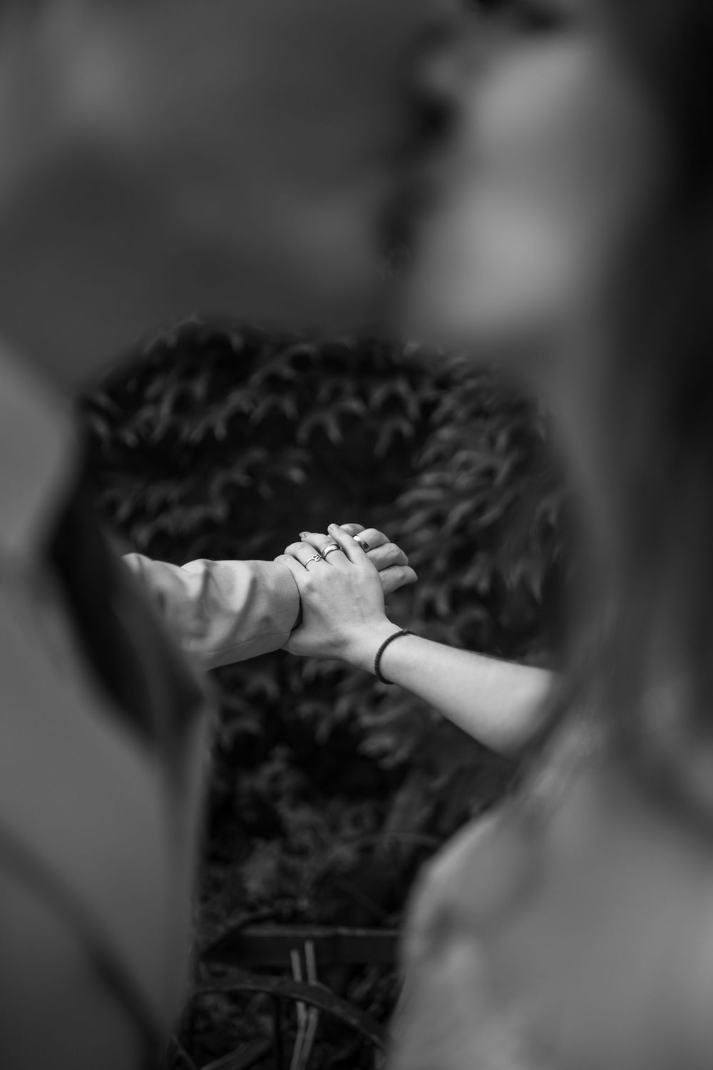 Black and white photo of a couple holding hands, with an engagement ring visible, set against a blurred natural background.