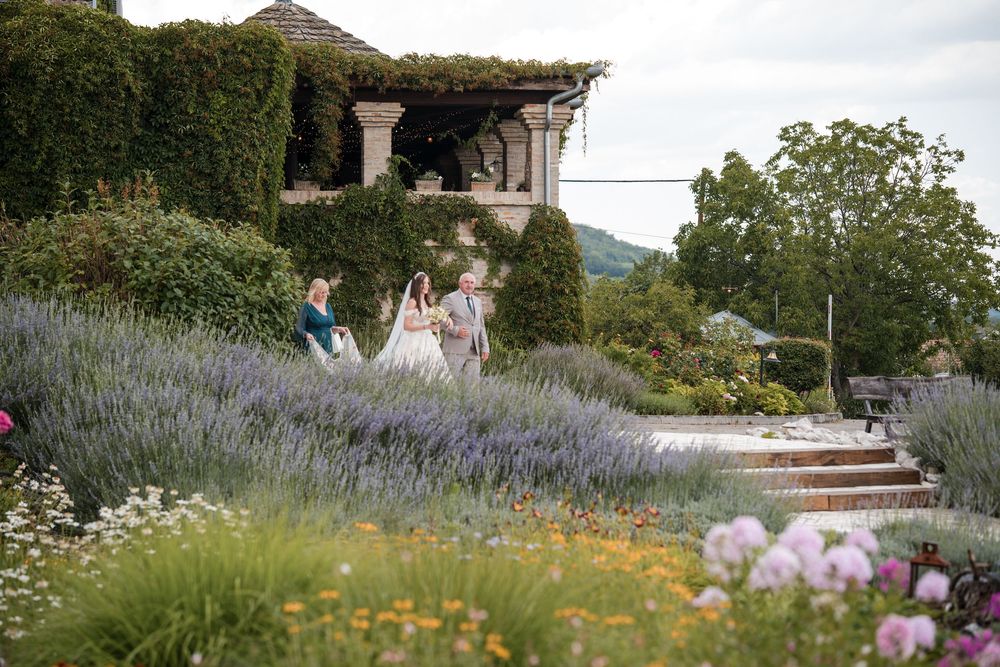 Wedding party walks through lush lavender garden with stone pergola and colorful wildflowers at scenic outdoor venue.