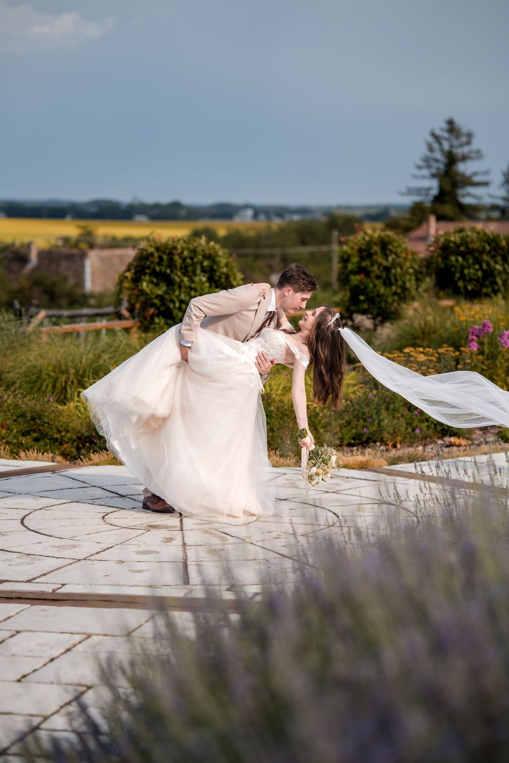 Groom dips bride for a romantic kiss in a garden setting, her veil flowing in the breeze on their wedding day.