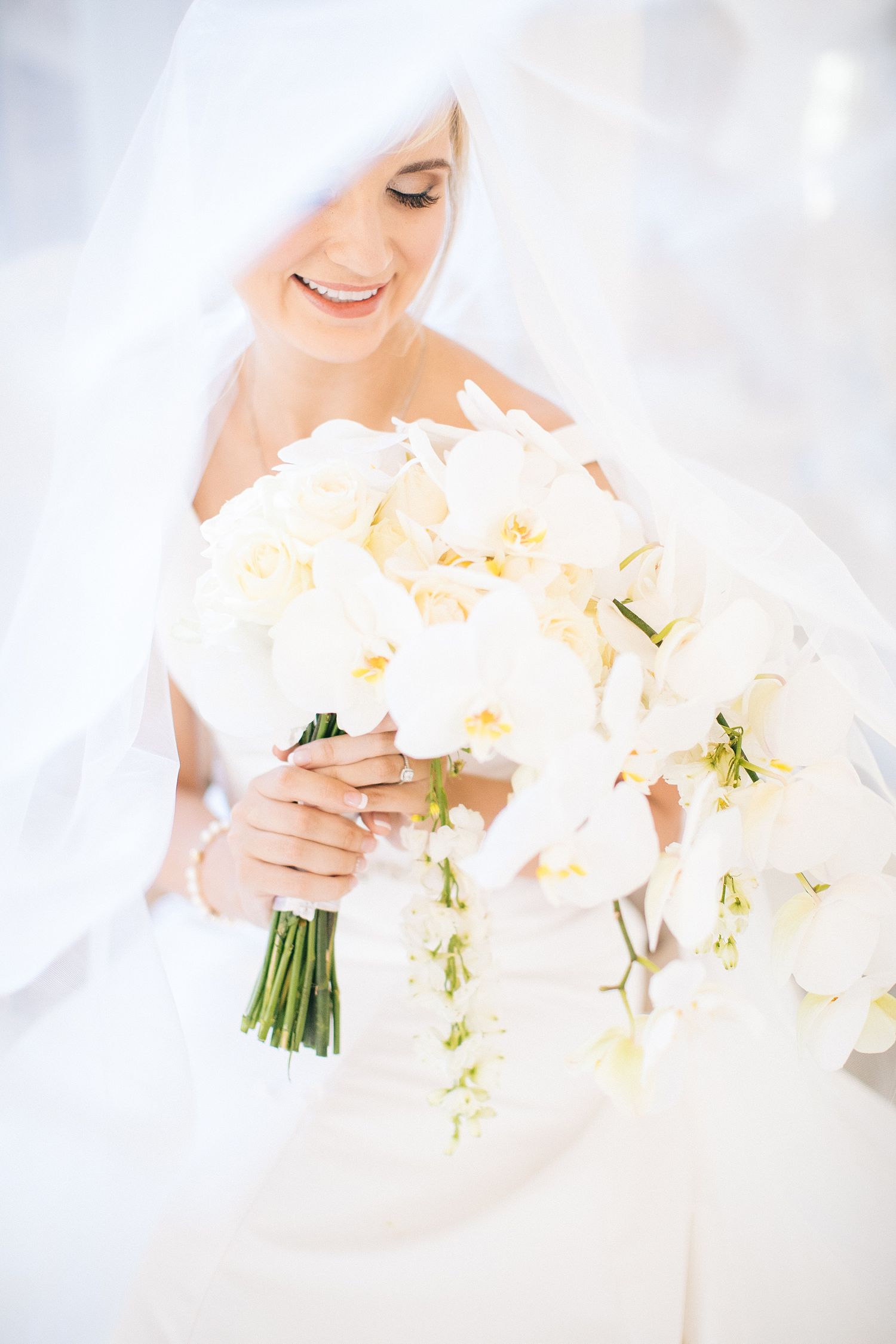 Smiling bride in white dress and veil holding a large bouquet of white orchids on her wedding day.