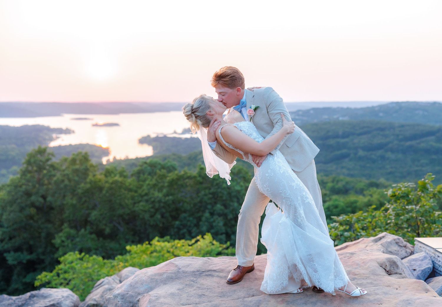 Bride and groom share a romantic dip kiss on a rocky overlook at sunset with a lake and forest below.