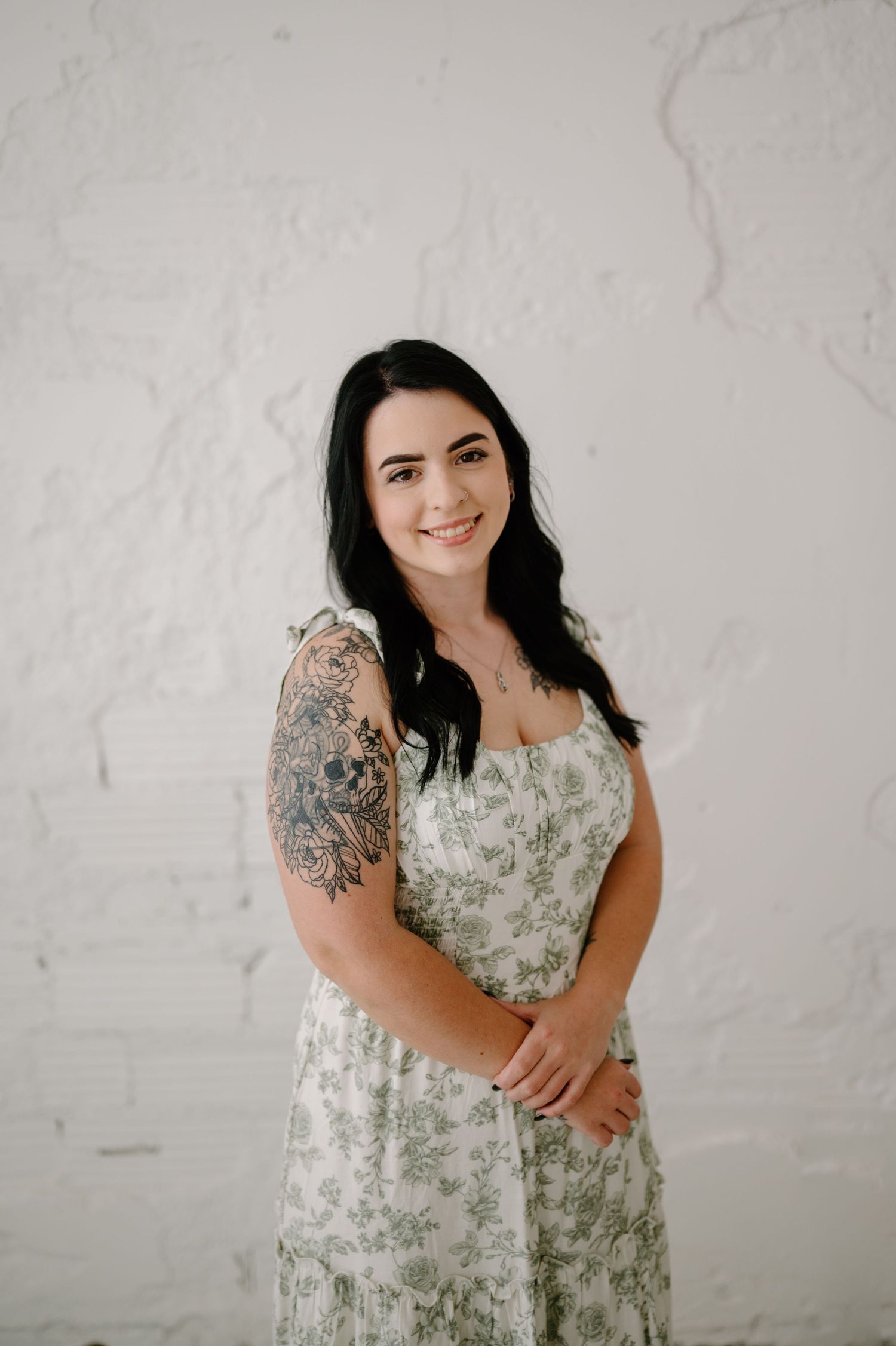 Woman in floral sundress with sleeve tattoo smiling against white brick wall.