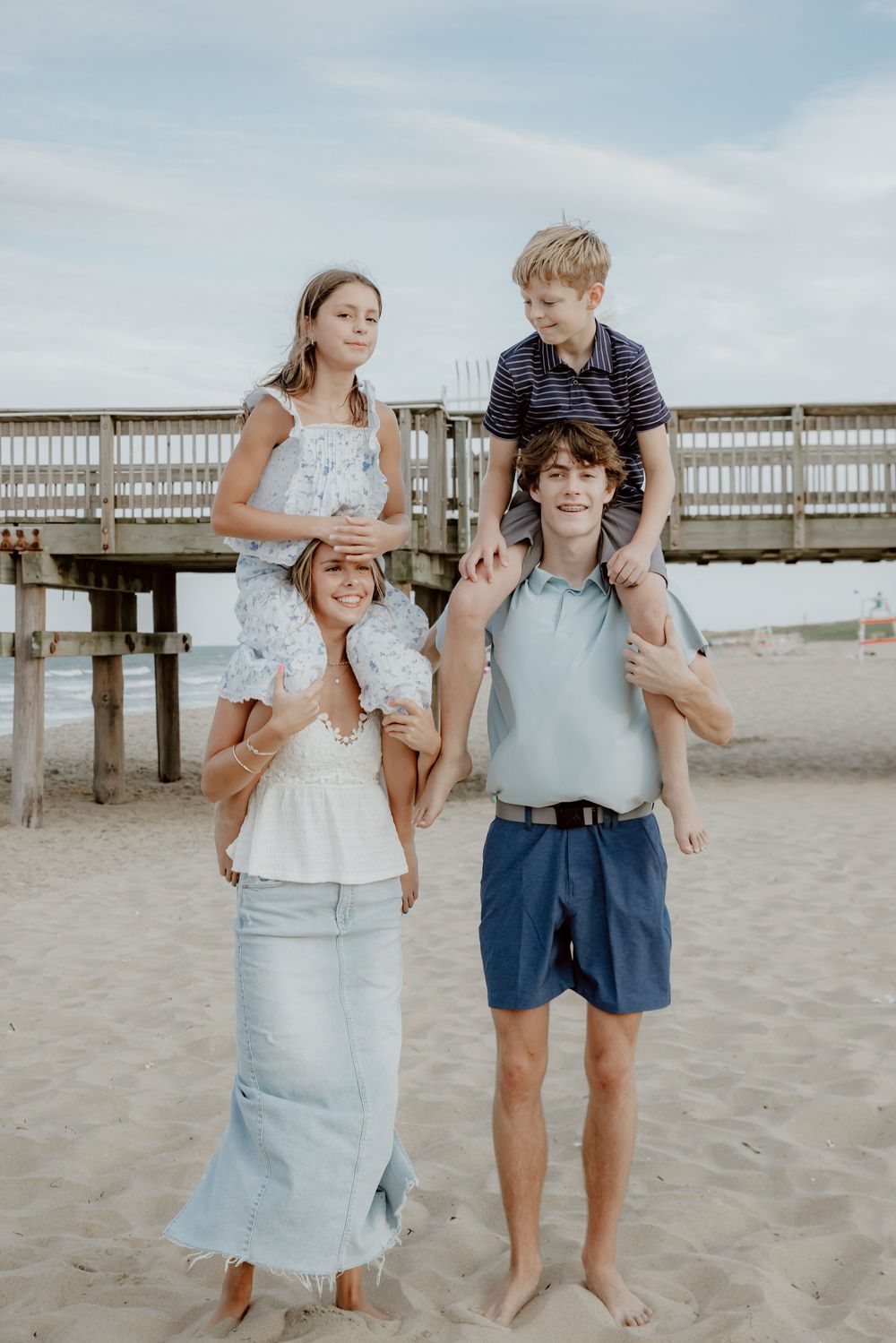 Family of four posing on sandy beach with pier backdrop, two kids riding on parents shoulders. Sandbridge.
