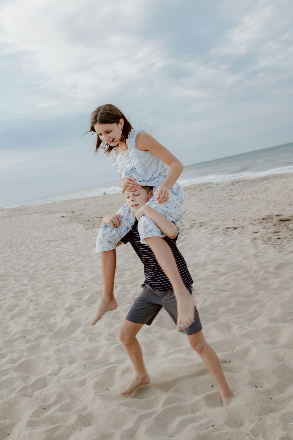 Young girl getting a piggyback ride from a boy on a sandy beach with ocean waves in background. Sandbridge.