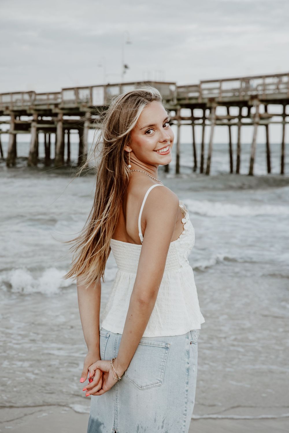 Smiling teenage girl with long blonde hair posing near a beach pier in white top and jeans. Sandbridge.
