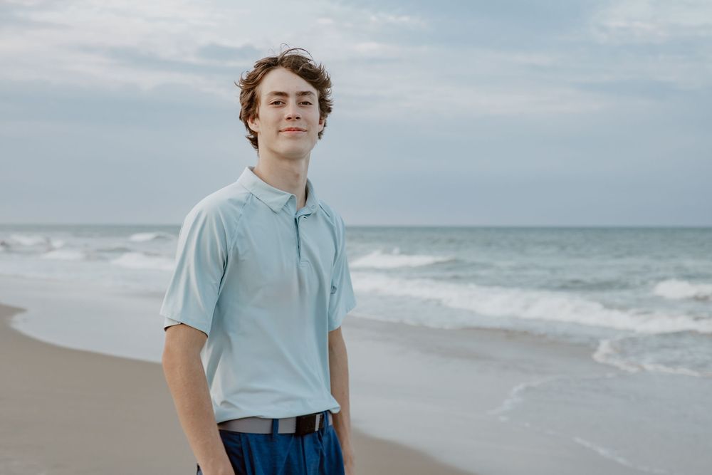 Teen boy in light blue polo shirt standing on beach with ocean waves behind him. Sandbridge.