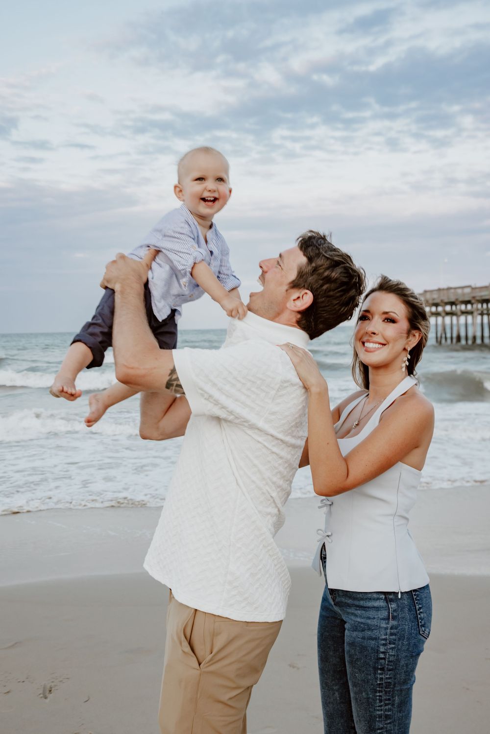 Happy couple lifting laughing toddler boy in the air on a beach near a wooden pier. Sandbridge.