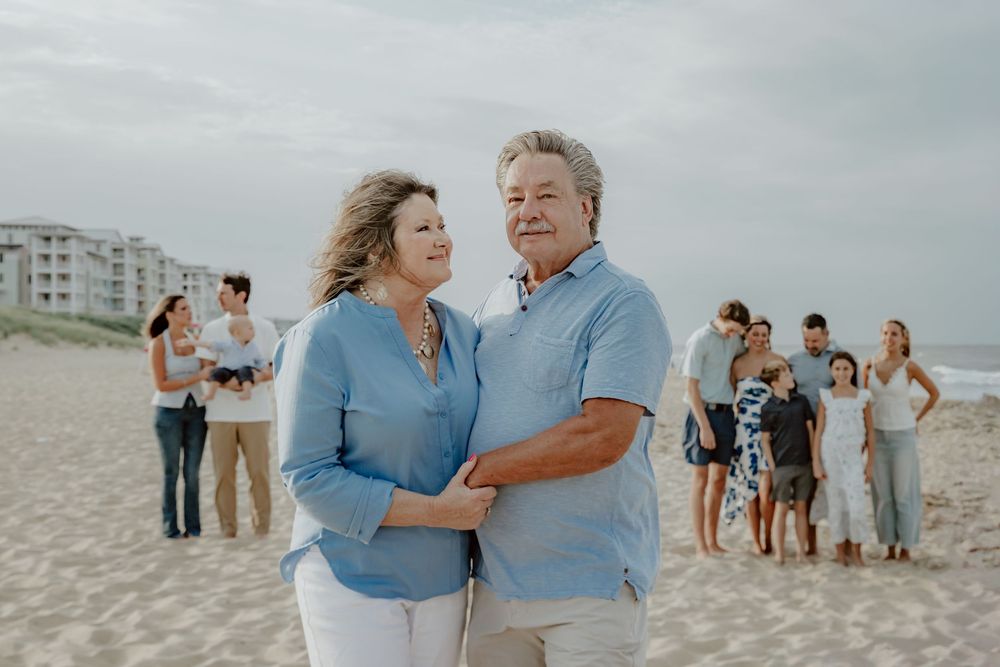 Smiling older couple in matching blue shirts embrace on sandy beach with family gathered behind them. Sandbridge.