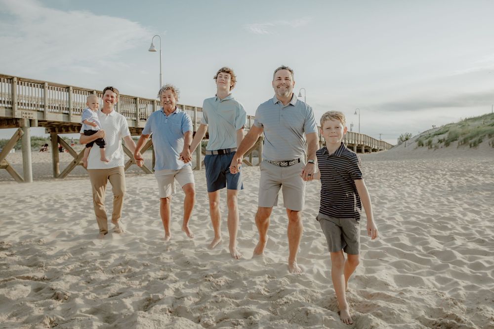 Group of five males of varying ages walk and jump together on sandy beach near wooden pier. Sandbridge.