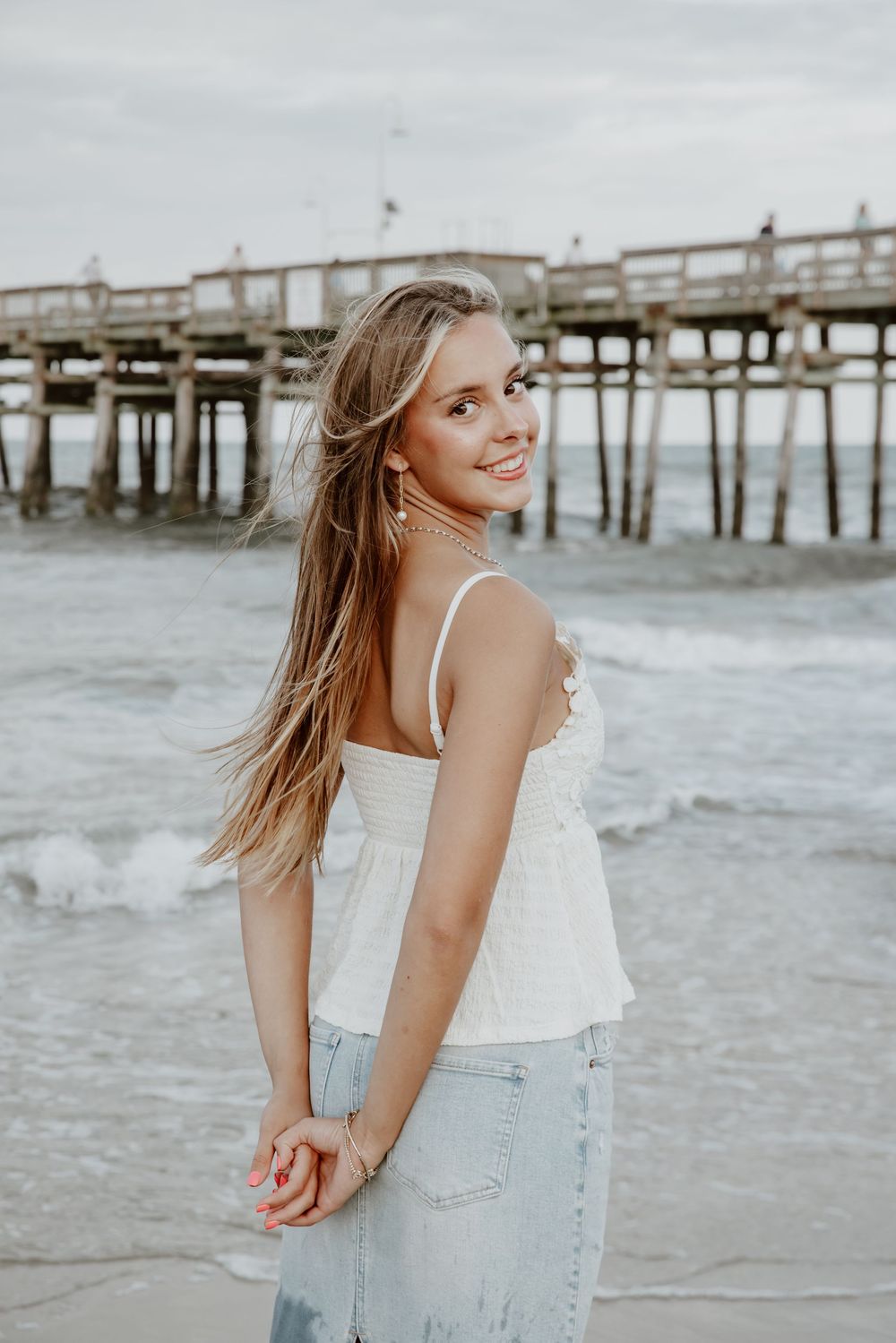 Young blonde woman in white top and jeans smiles over shoulder near ocean pier at sunset. Sandbridge.
