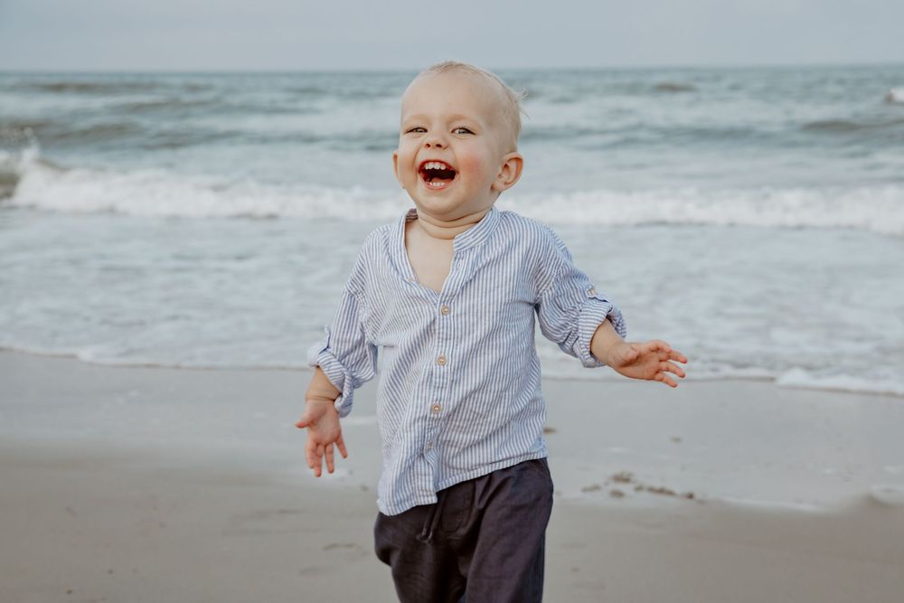 Laughing toddler boy running on the beach wearing a blue button-up shirt with ocean waves behind him. Sandbridge.