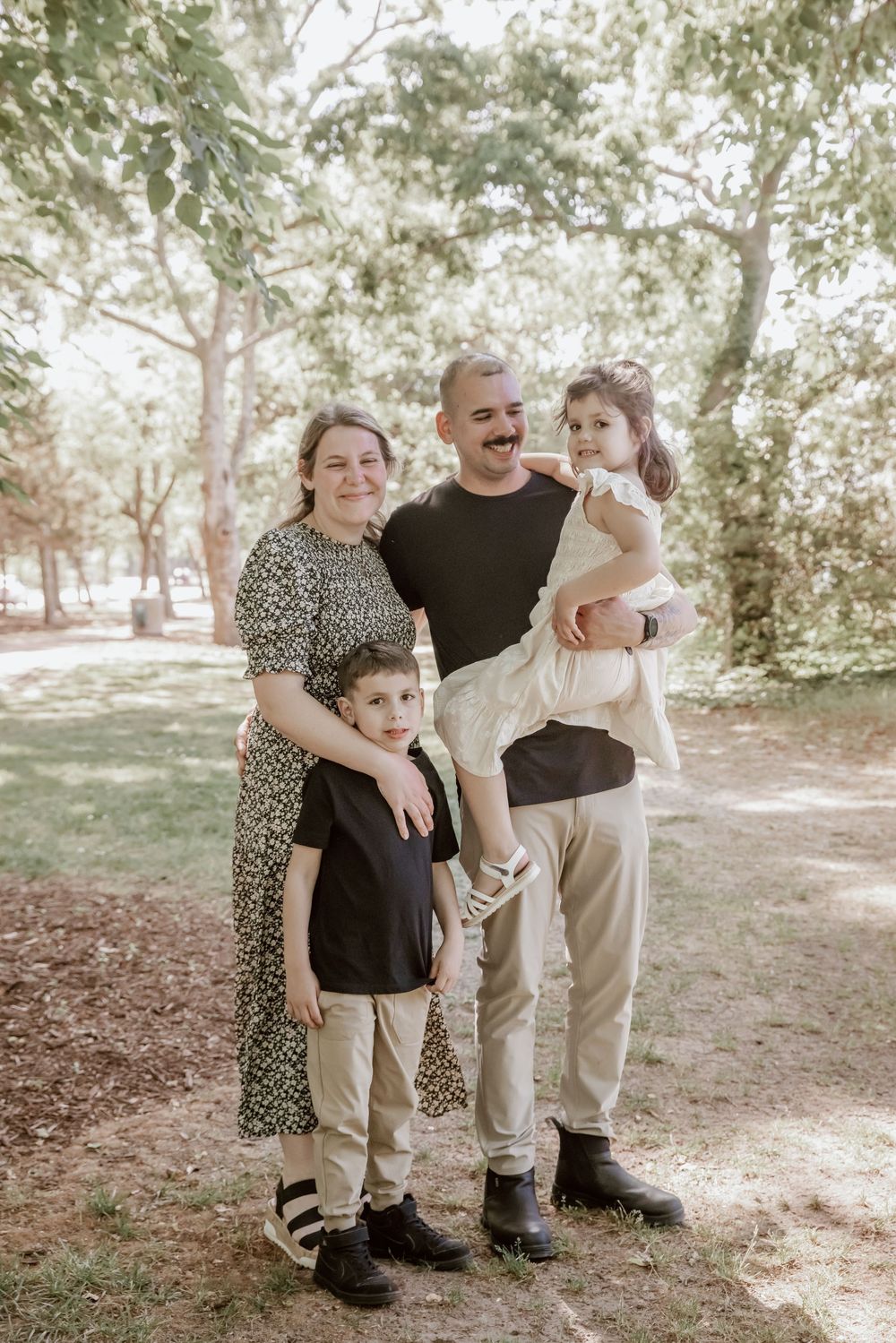 Smiling family of four stands together among trees during casual outdoor park photo session. Yorktown.