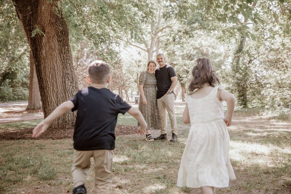 Two children run toward parents standing near large tree during playful outdoor family photo session. Yorktown.