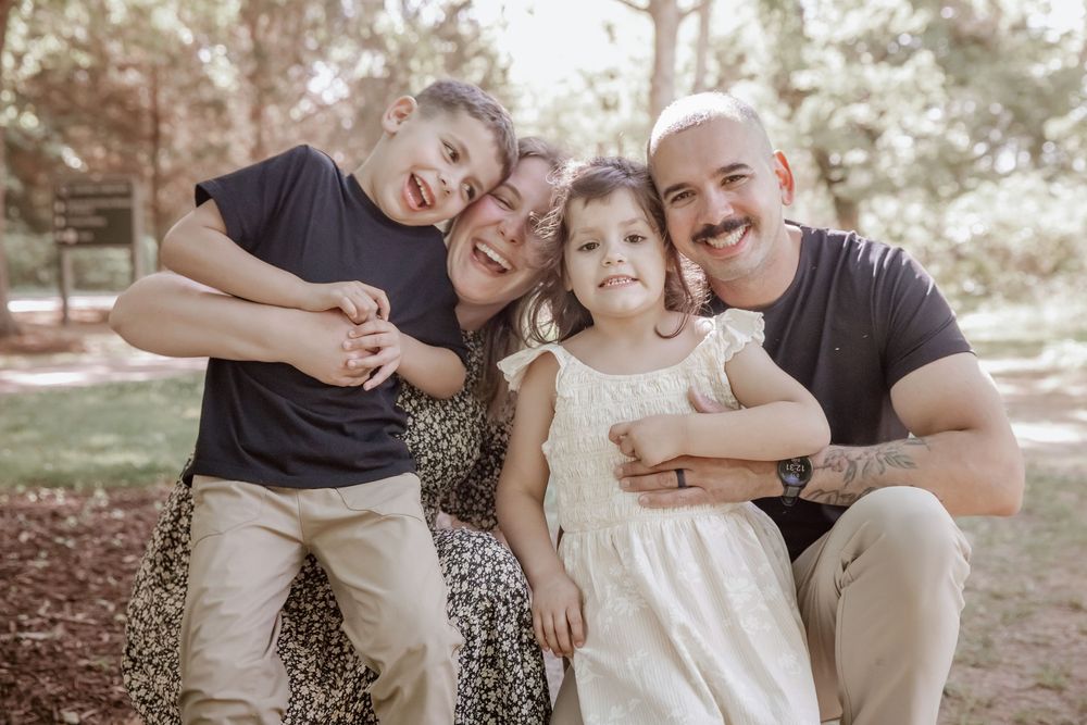 Joyful family of four cuddles close together for candid outdoor portrait in sun-dappled park setting. Yorktown.
