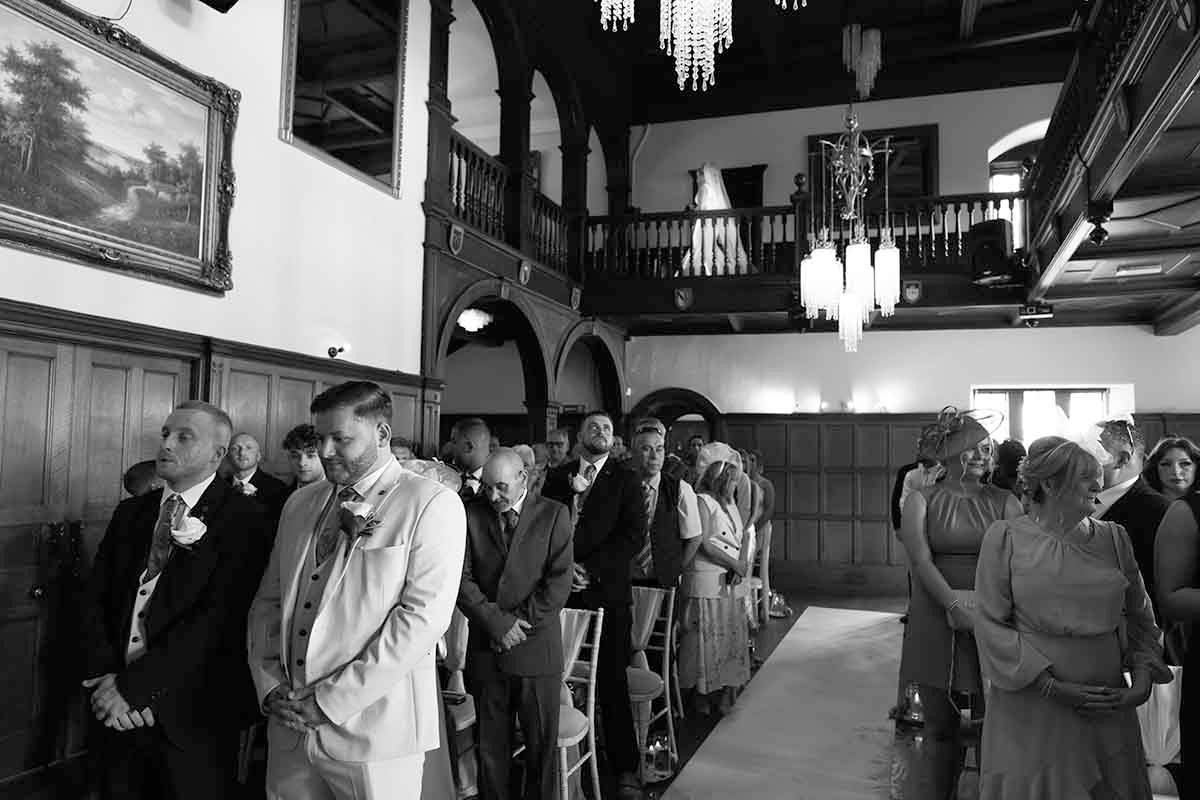 Groom waits at the altar as his bride passes above on the stairs at Dalston Hall wedding venue