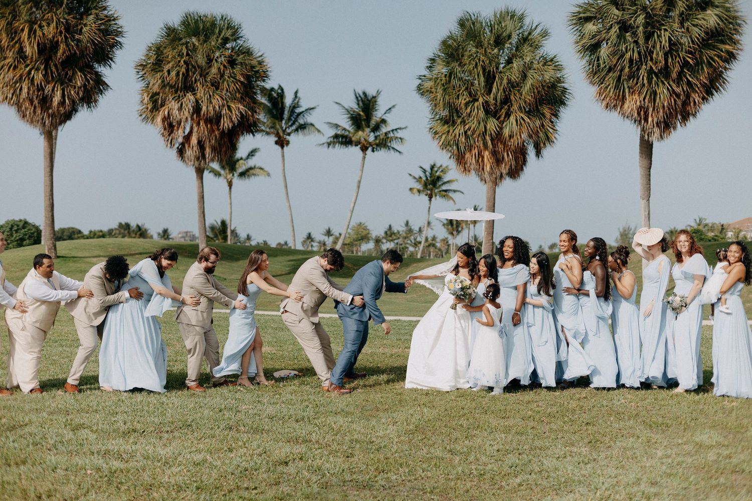 Wedding party in light blue and tan attire celebrating outdoors on a green lawn with tall palm trees in Florida.