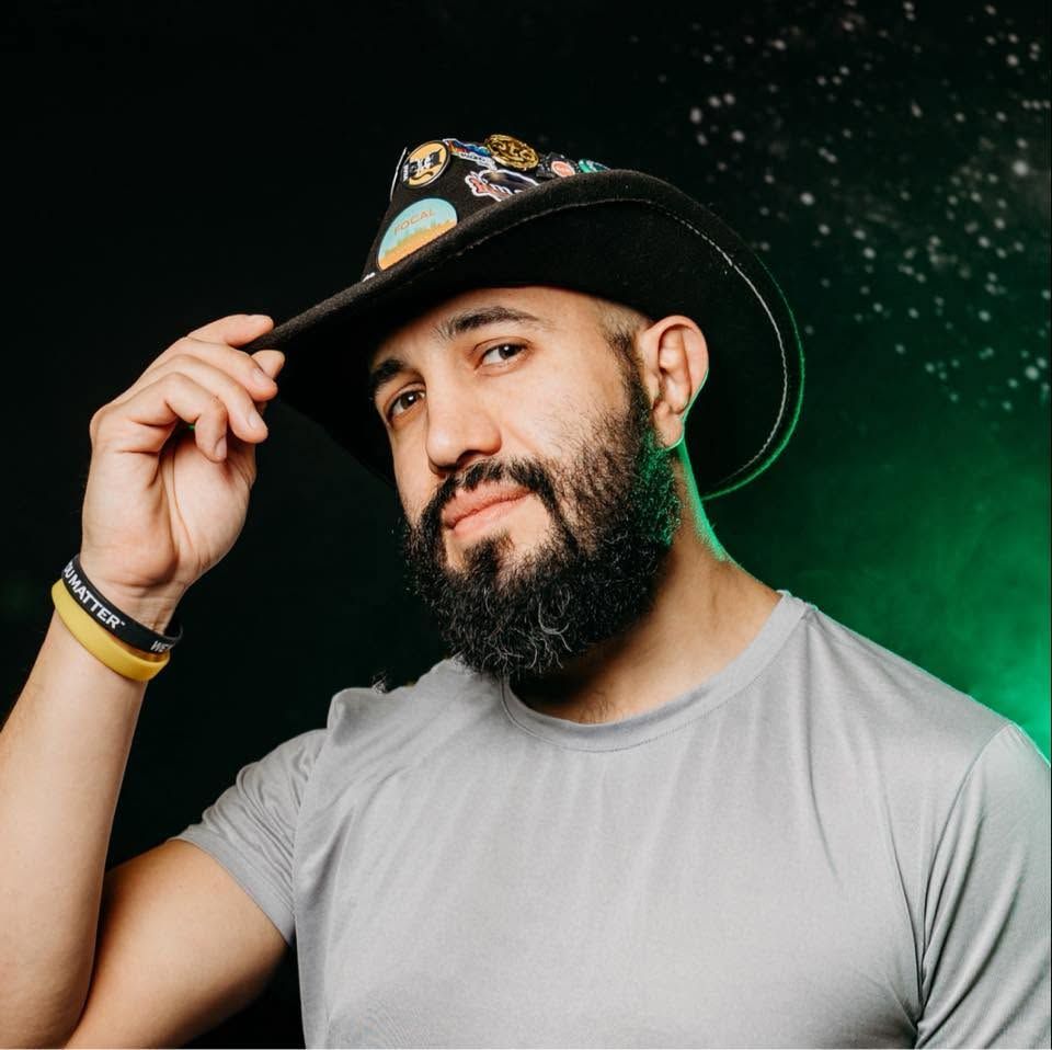 Bearded man in gray t-shirt and black cap with pins poses against dark green bokeh background.