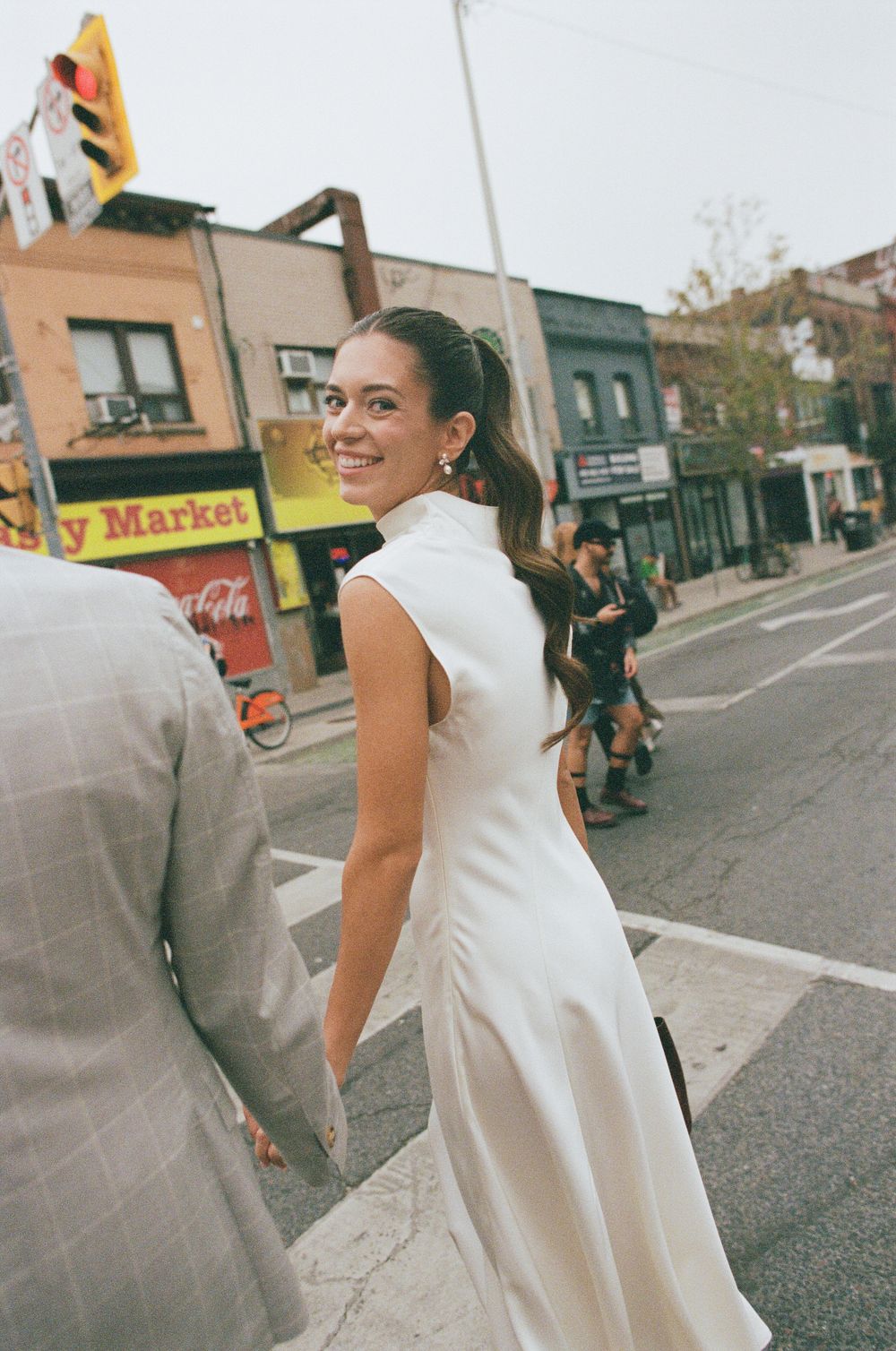 Smiling bride in white gown crosses urban street, glancing back, with shops and pedestrians in the background.
