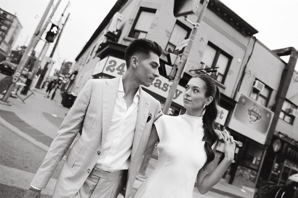 Black and white photo of an elegant couple in white outfits sharing a romantic moment on a city street corner.