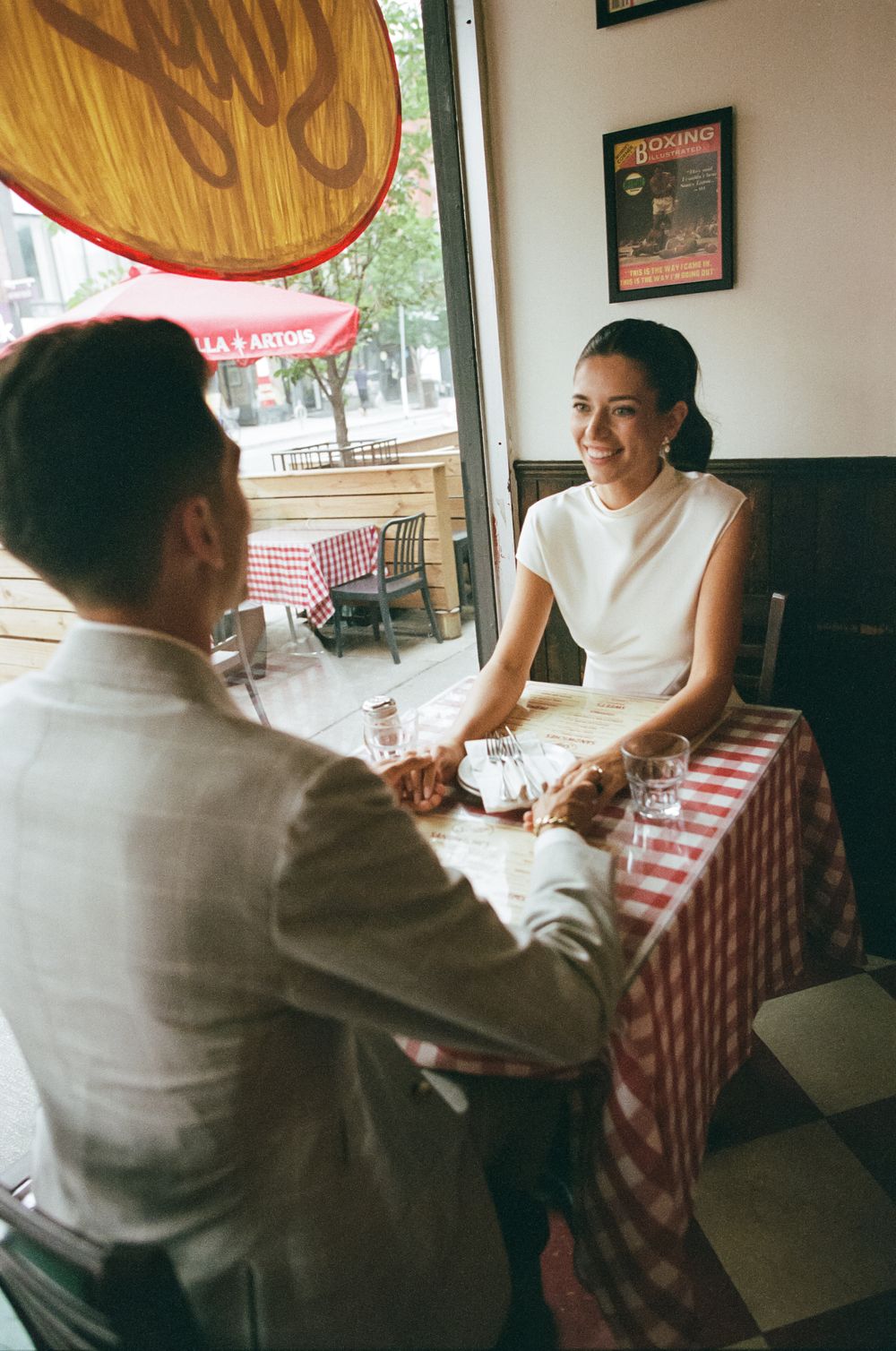 Couple sharing a romantic dinner at a cozy Italian restaurant with red checkered tablecloth and large window view.