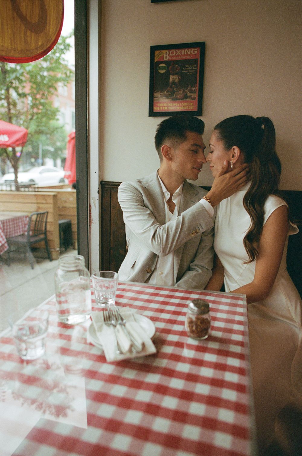 Couple sharing a romantic moment at a red checkered tablecloth restaurant, dressed in elegant white wedding attire.