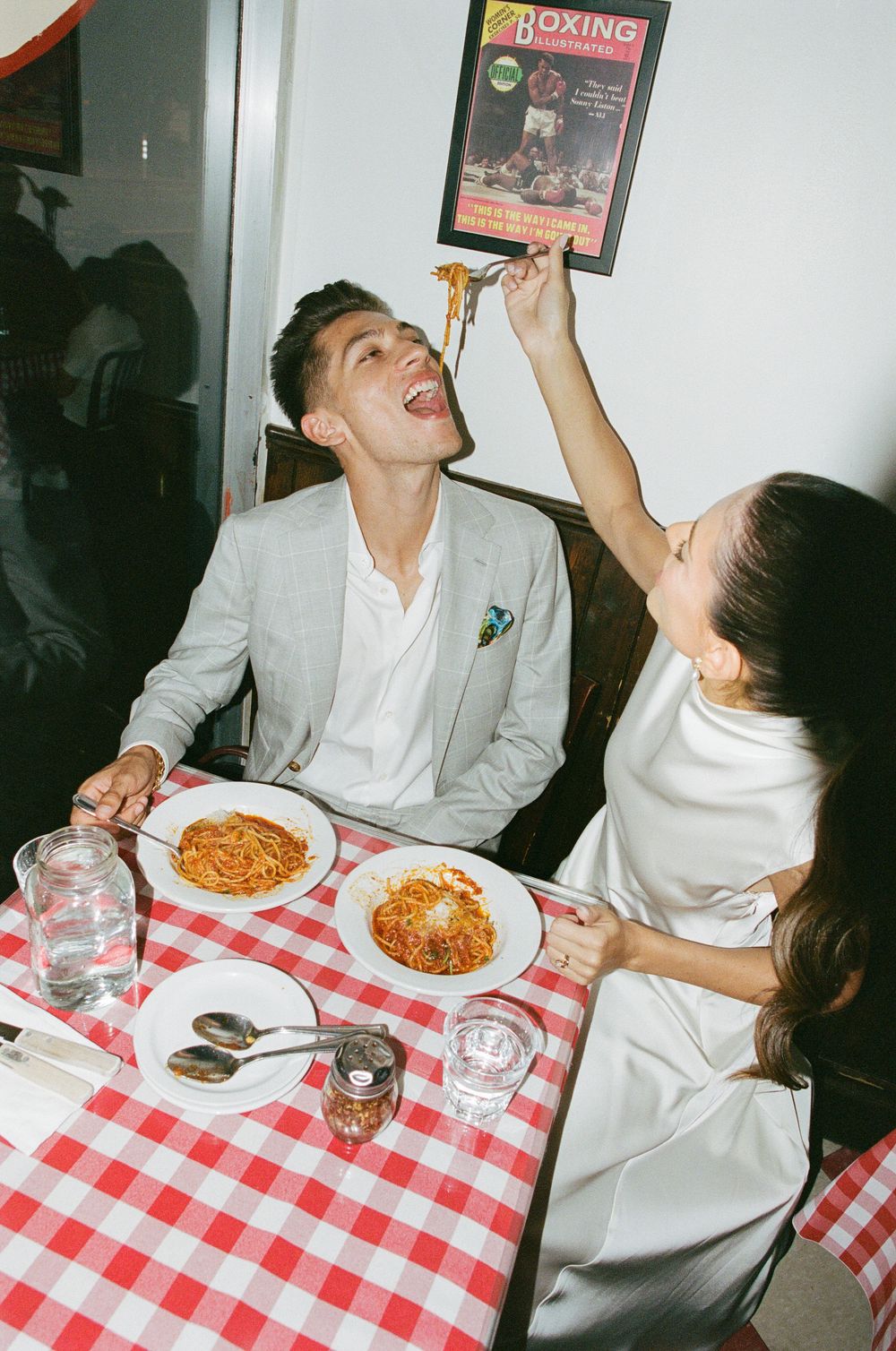 Couple sharing spaghetti at a red checkered tablecloth restaurant, woman feeding man pasta playfully.