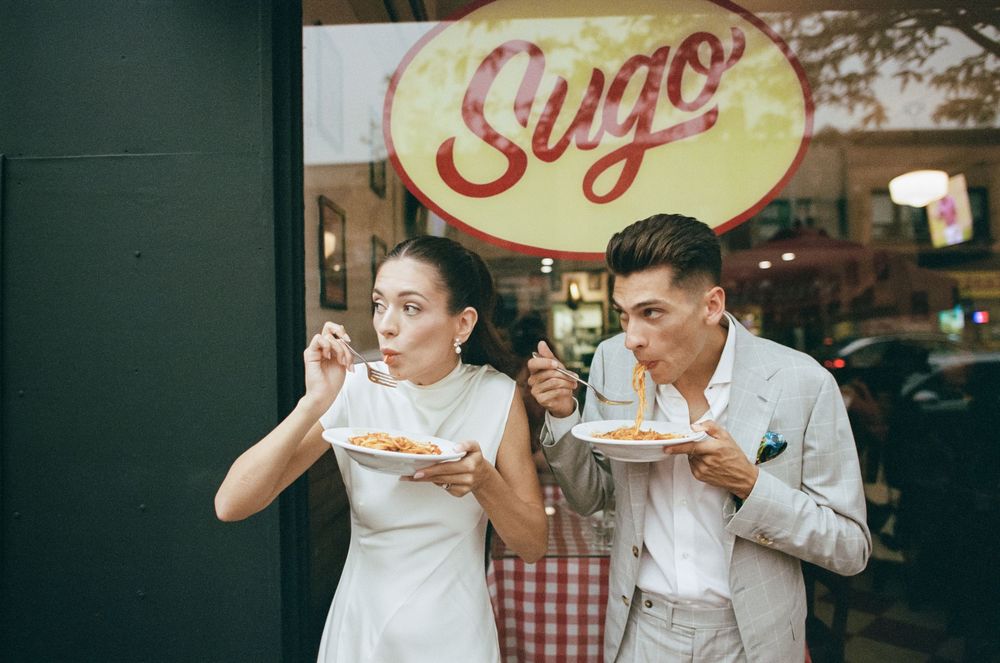 Couple eating pasta outside Sugo restaurant, woman in white dress and man in gray suit sharing Italian food.