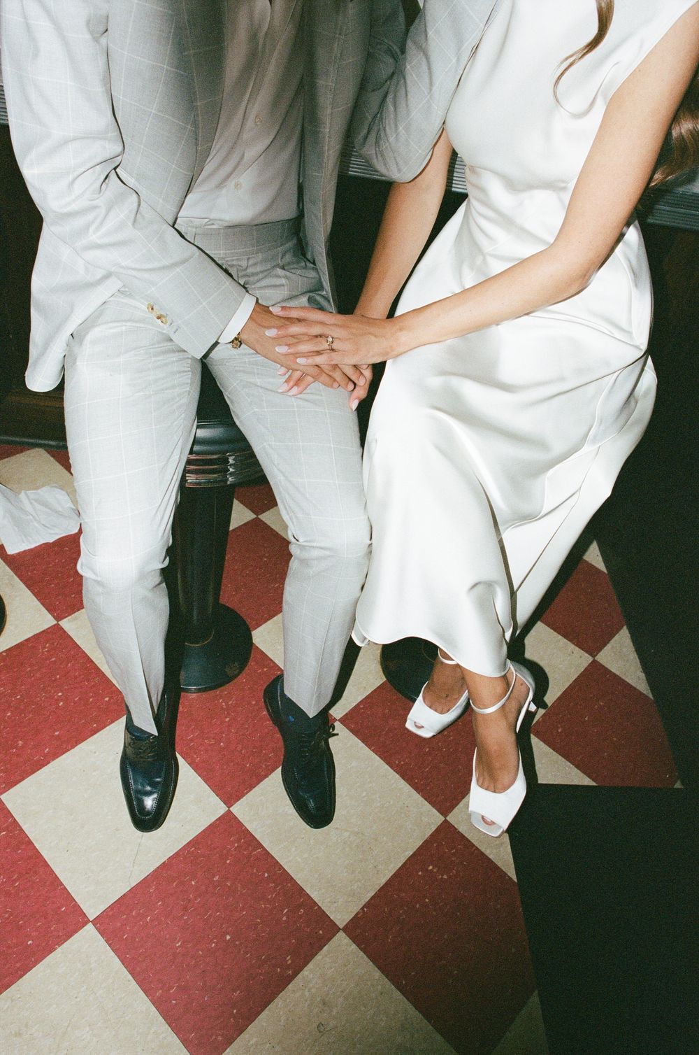 Bride and groom sitting together holding hands, wearing white wedding attire on a red checkered floor.