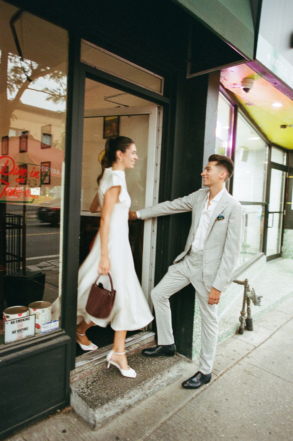 Elegant couple in wedding attire exiting a charming urban storefront on a city sidewalk.