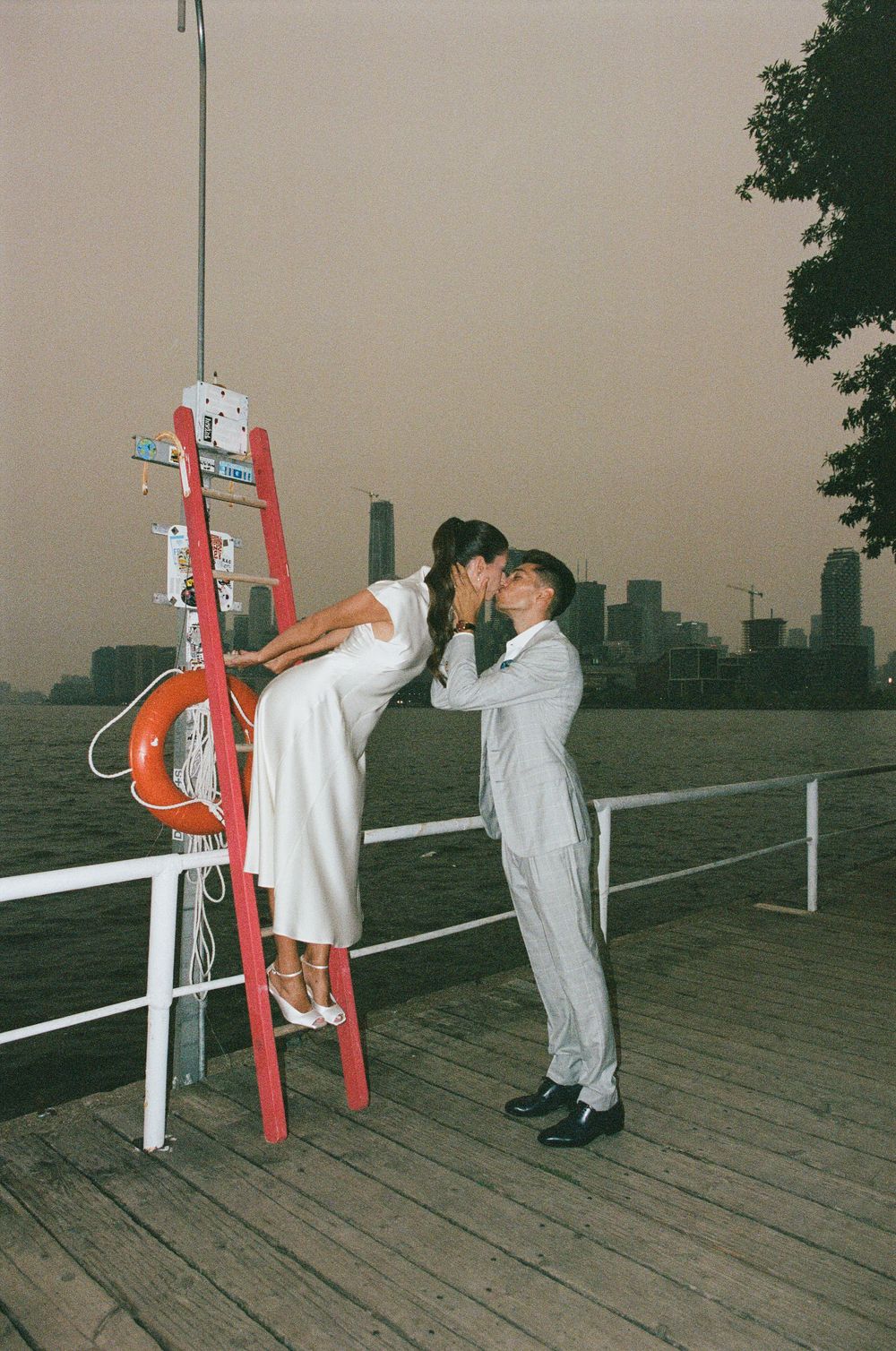 Couple sharing a romantic kiss on a wooden pier, woman on red ladder, city skyline at dusk in background.