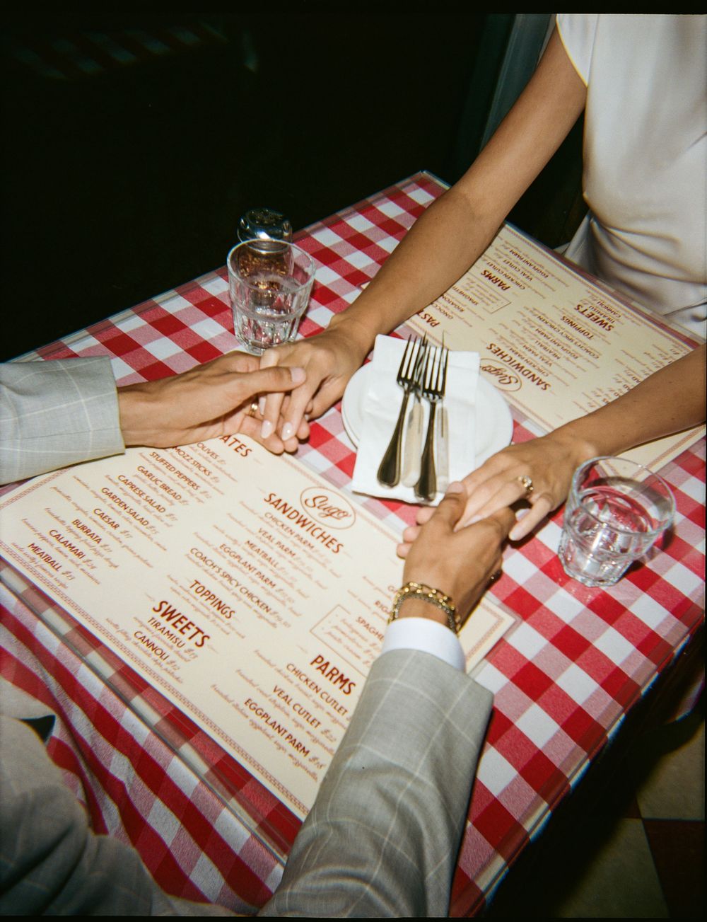 Couple holding hands at a red checkered tablecloth restaurant, with menus, water glasses, and silverware on table.
