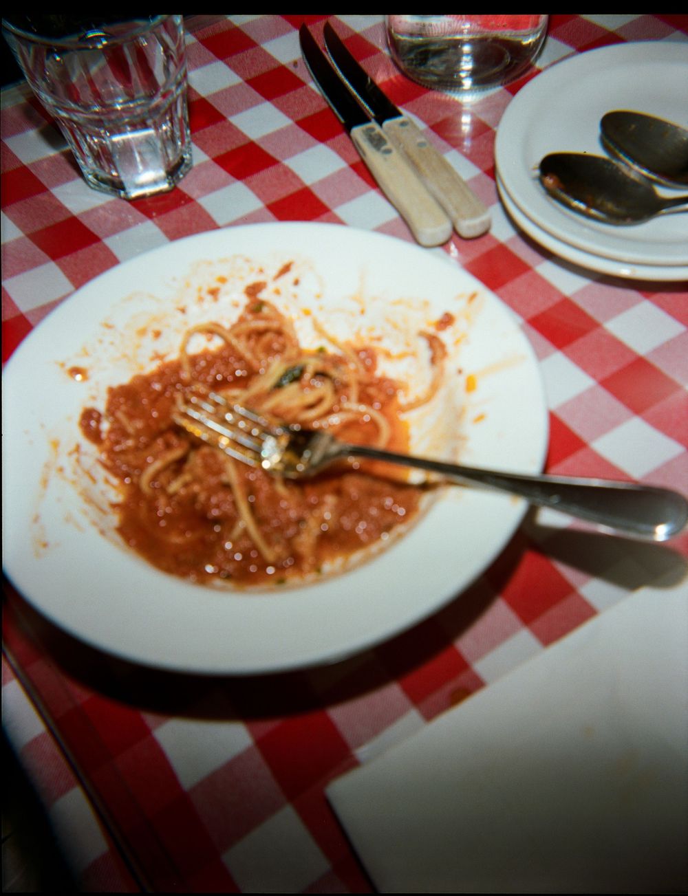 Half-eaten spaghetti with tomato sauce on white plate with fork, on red checkered tablecloth in Italian restaurant.