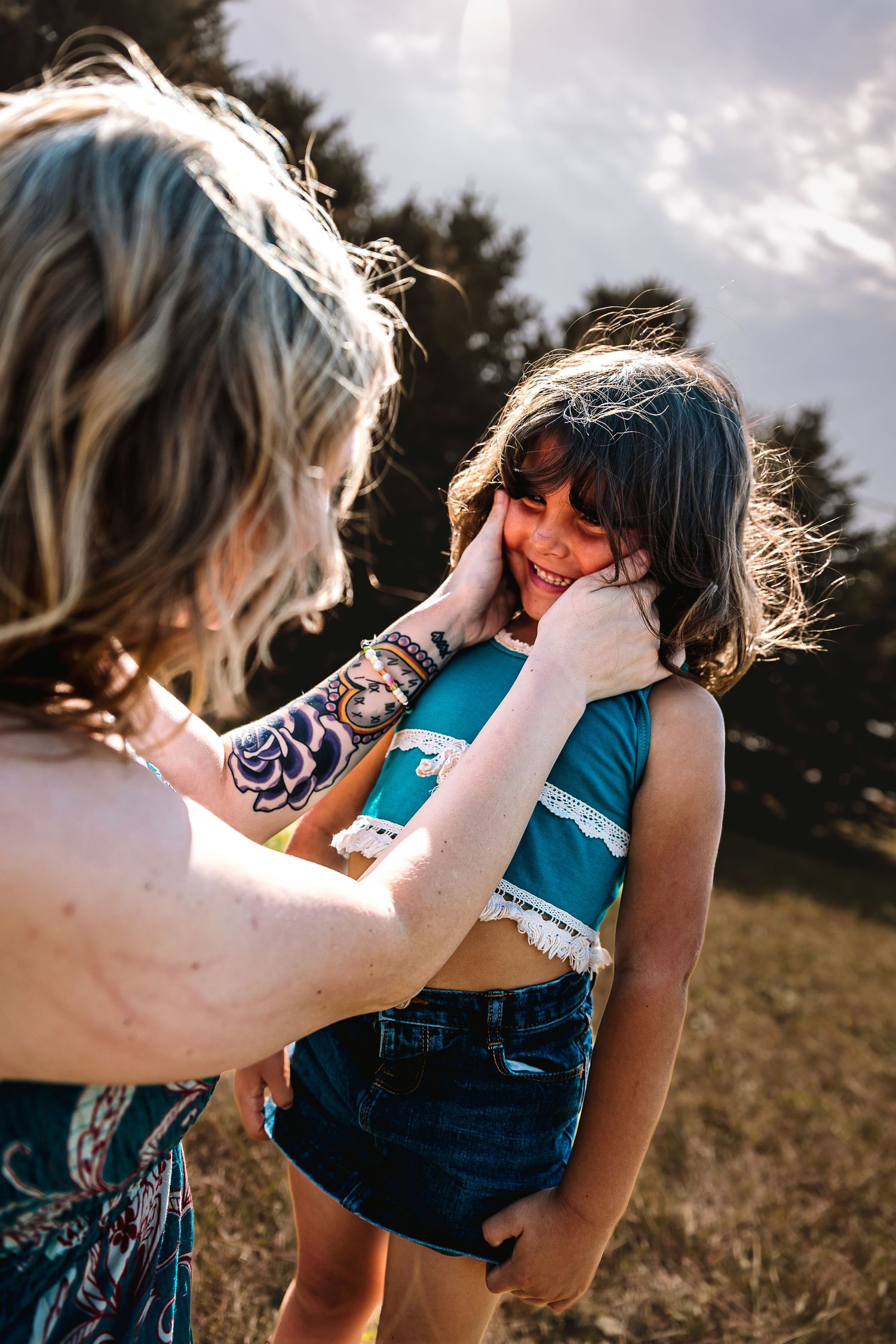 A smiling blonde woman in a floral dress hugs a laughing young girl outdoors in a sunny field.