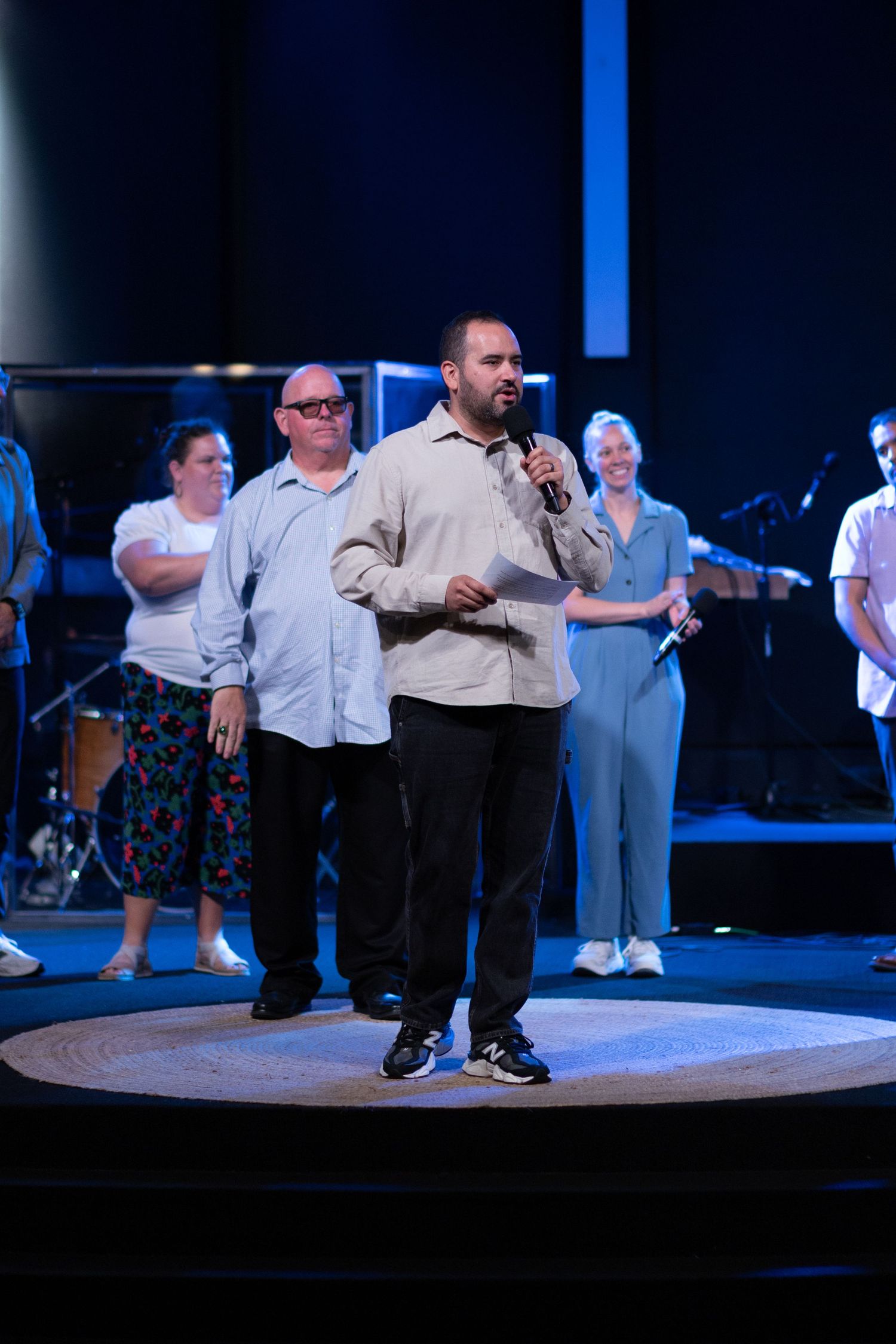 Man speaking into microphone on stage with group of people standing behind him in a dimly lit venue.