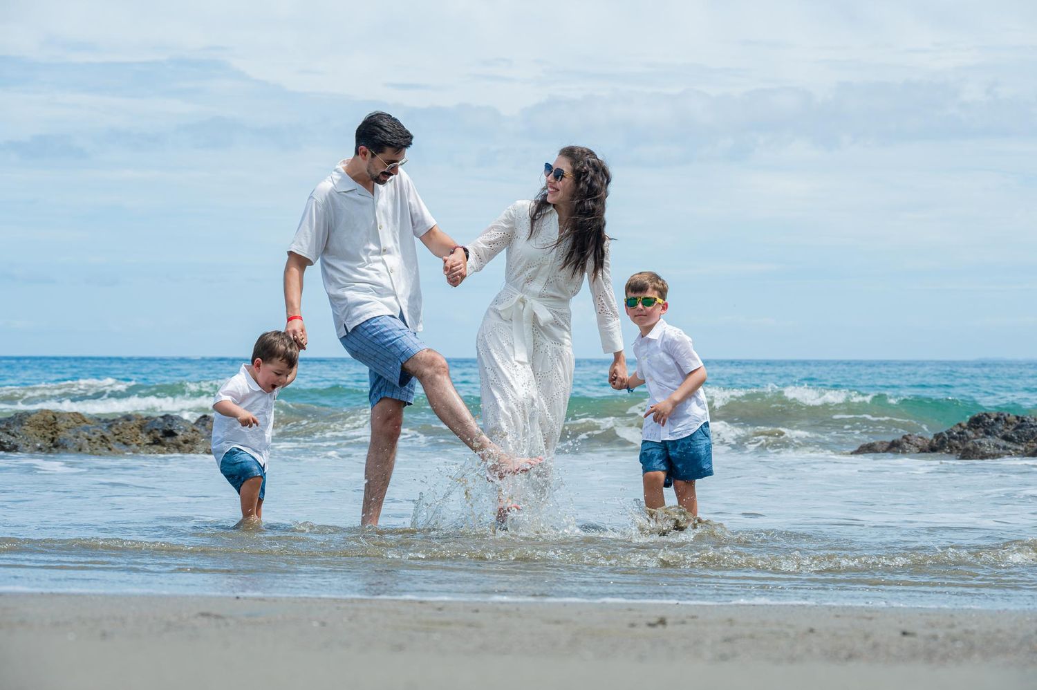 Family of four playing in ocean waves on a sunny beach, parents and two young children splashing in white outfits.