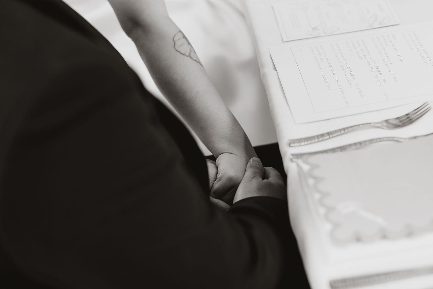 Black and white photo of a couple holding hands at a signing table during a wedding ceremony.