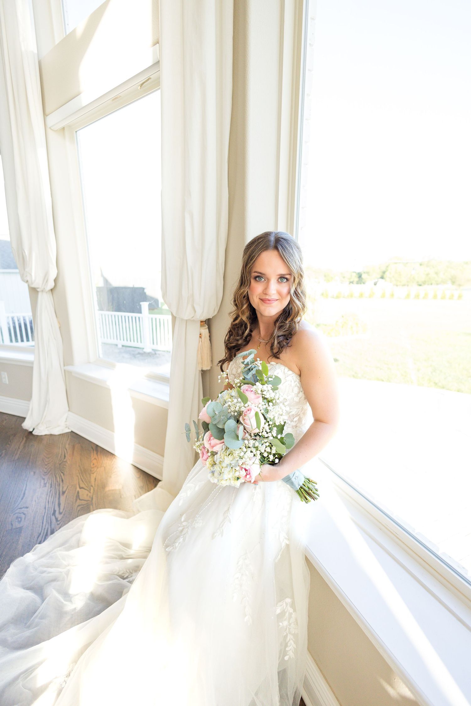 Bride holding a colorful wedding bouquet beside a bright window, wearing a flowing white gown during her bridal portrait session.