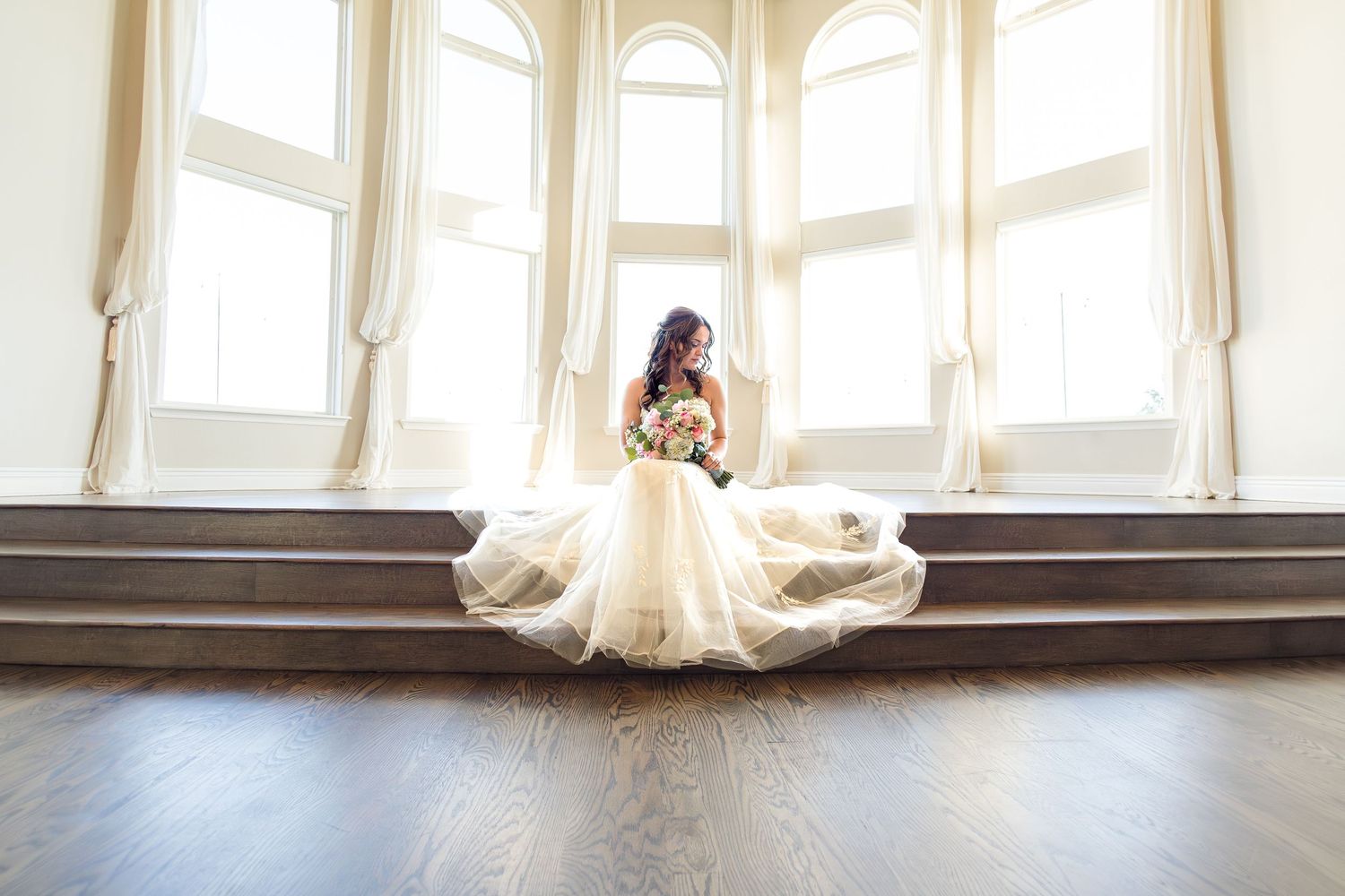 Bride sitting on wooden steps in a flowing wedding dress beneath tall windows during an elegant indoor bridal portrait session.