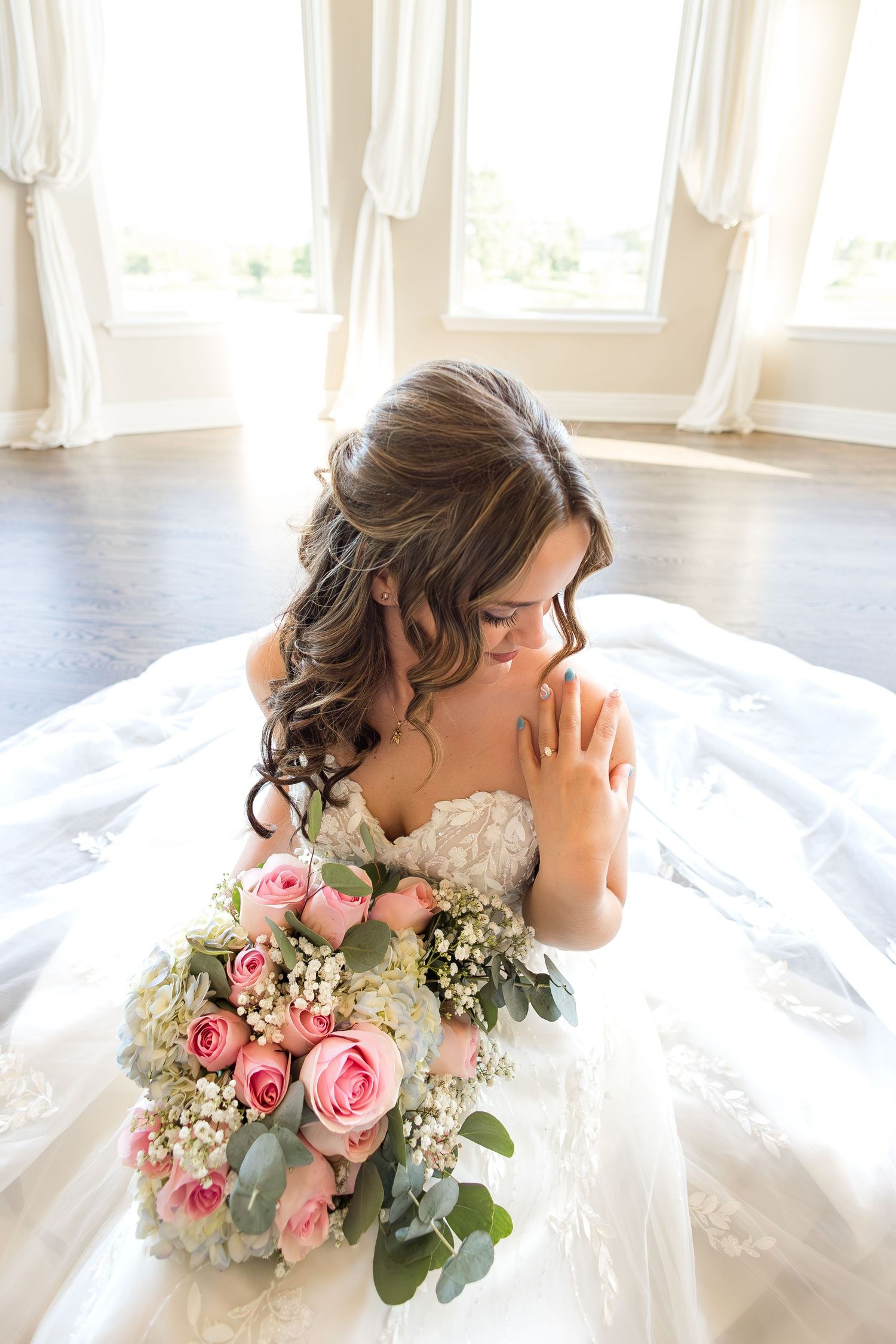 Bride seated indoors in a flowing white wedding gown, holding a colorful bouquet of pink flowers in soft natural light.
