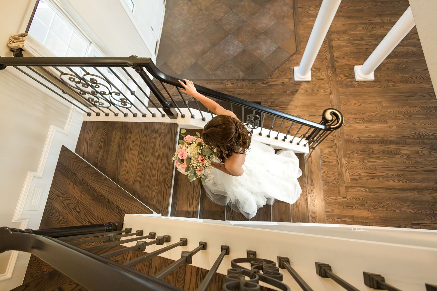 Overhead view of a bride in a white wedding gown holding her bouquet while standing on an elegant wooden staircase.