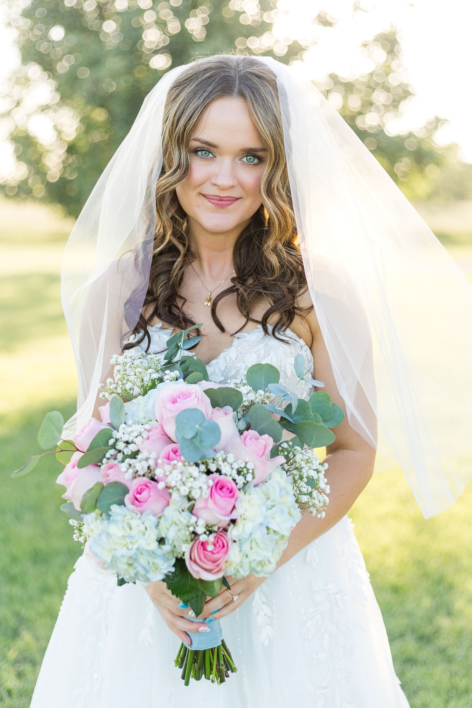 Smiling bride wearing a delicate wedding veil and holding a colorful bouquet of pink and white flowers outdoors.