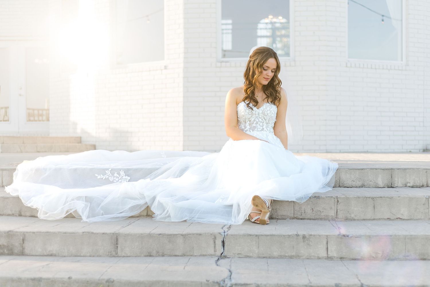 Bride in a flowing white wedding gown sitting on outdoor stone steps during a bright, romantic bridal portrait session.