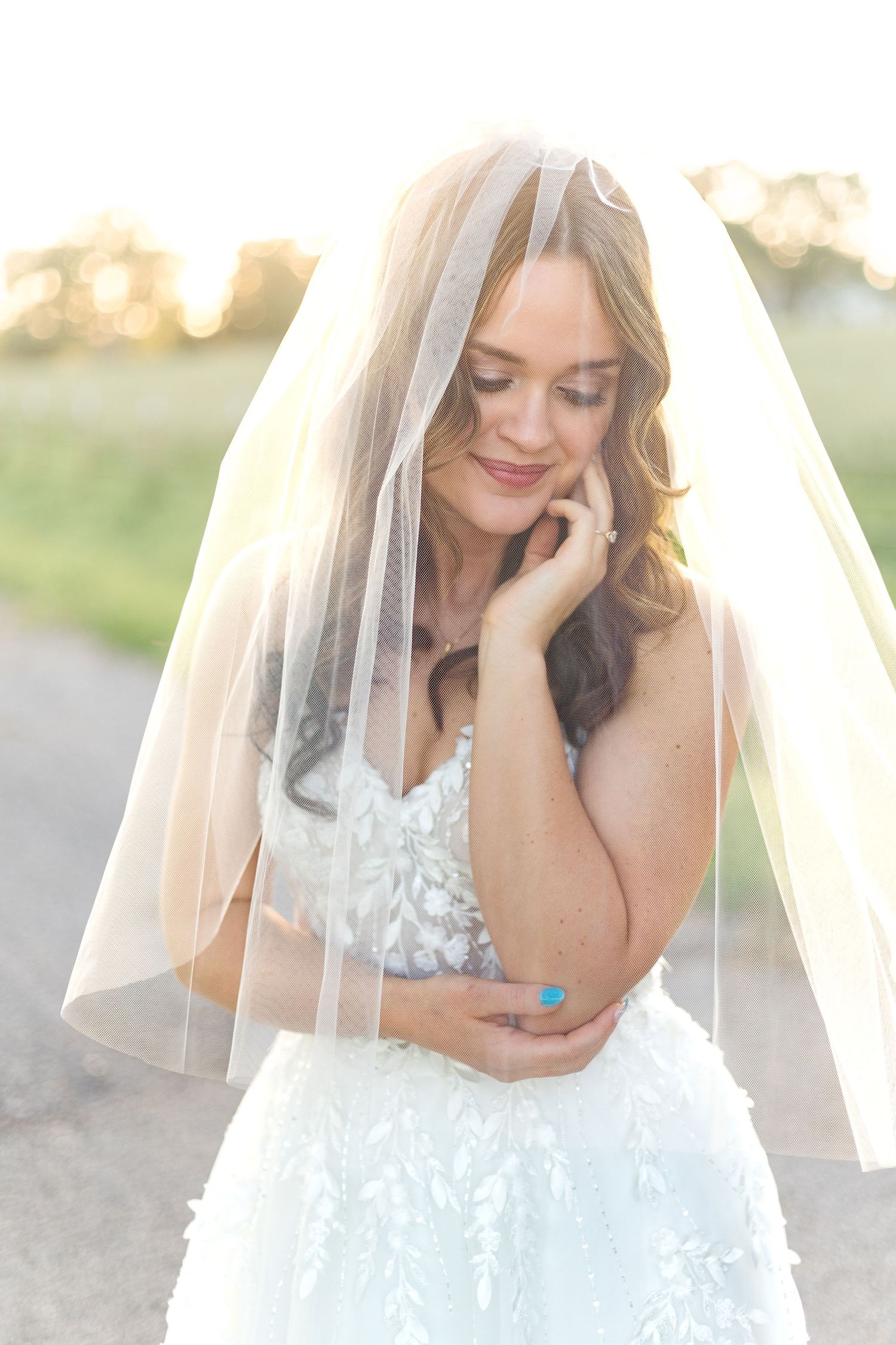 Bride smiling in a white lace wedding gown and delicate veil during a romantic outdoor bridal portrait session.